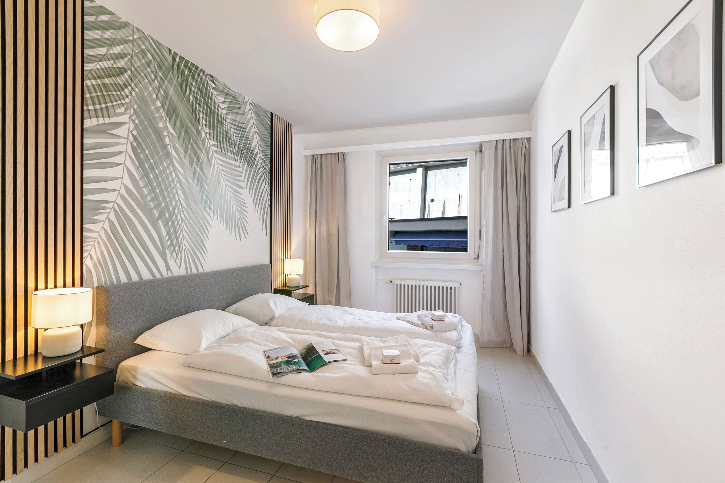 Ein modernes Hotelzimmer mit einem großen Bett, Nachttischlampen, einem Fenster mit Vorhängen und gerahmten Bildern an der Wand.