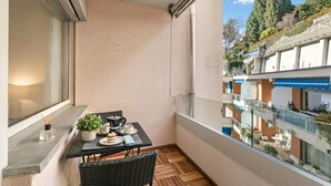 A balcony with a table set for two, a potted plant, and a window with a view of a building and trees. - Residenza Tasso Sul Lago in Castagnola (Lugano)