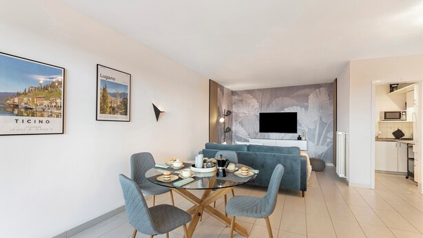 A modern living room with a glass dining table, grey upholstered chairs, a blue sofa, a flat-screen TV, and a kitchen area. - Residenza Tasso Sul Lago in Castagnola (Lugano)