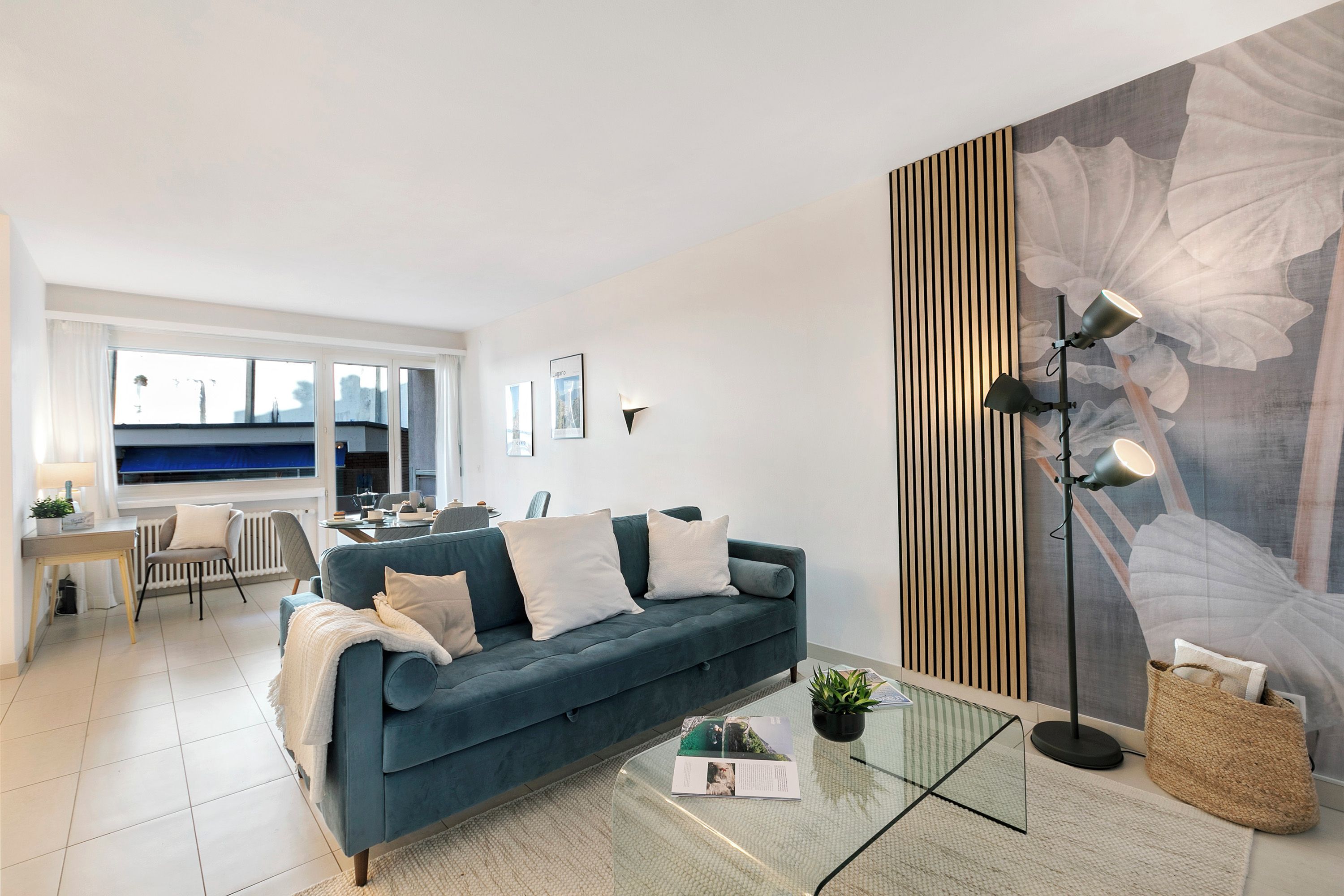 Una sala de estar moderna con un sofá azul, una mesa de centro de vidrio, una lámpara de pie y un mural floral grande en la pared.