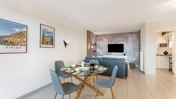 A modern living room with a glass dining table, grey upholstered chairs, a blue sofa, a flat-screen TV, and a kitchen area.