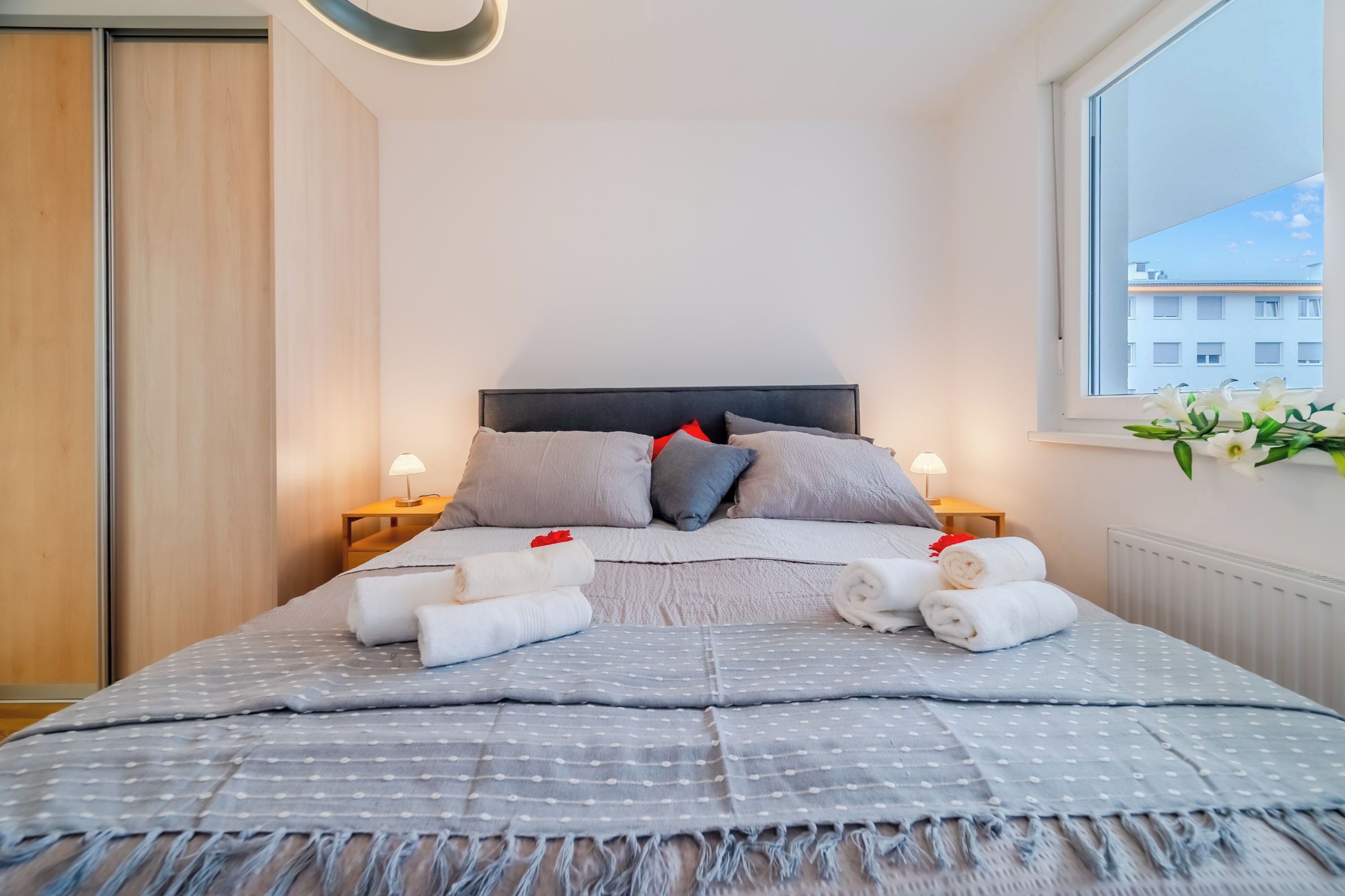 Ein ordentlich gemachtes Bett mit Kissen und Handtüchern, eine Nachttischlampe und ein Fenster mit Blick auf Gebäude.