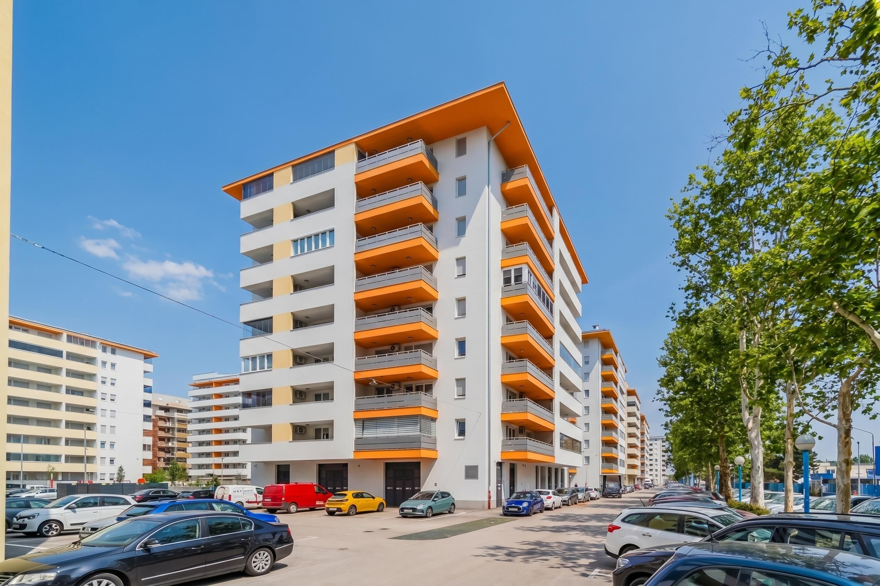 A multi-story residential building with balconies, parked cars, and trees along the street.