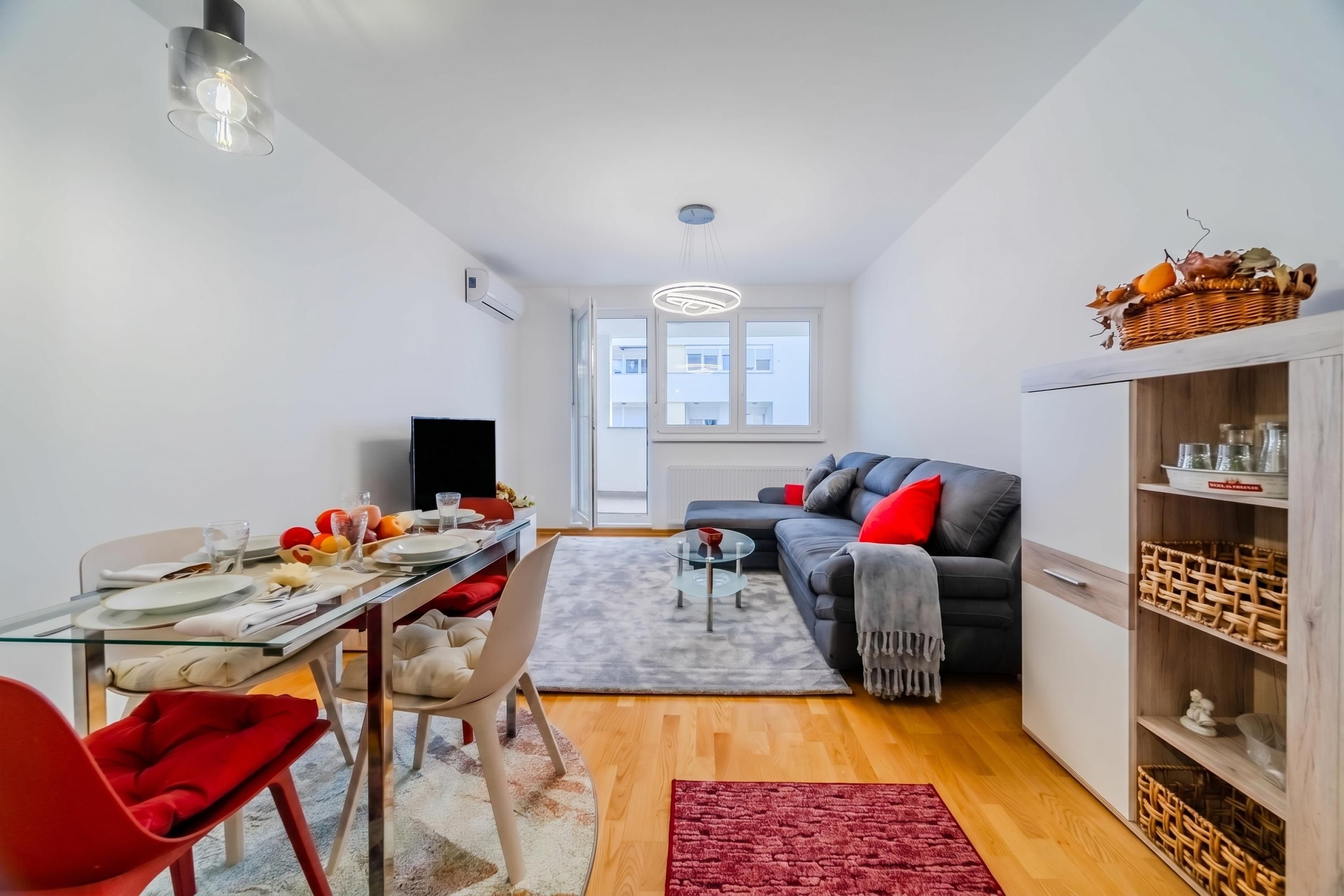 A modern living room with a dining area, a grey sofa, a red chair, a glass coffee table, and a red rug.