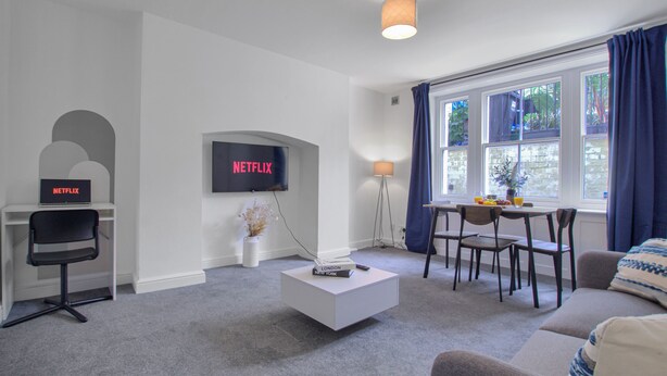 Un salon moderne avec un téléviseur à écran plat fixé au mur, une table basse blanche, un canapé, une table à manger avec des chaises et une fenêtre ornée de rideaux bleus.