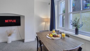 Un coin repas avec une table mise pour le petit-déjeuner, un écran de télévision affichant le logo « NETFLIX », et une fenêtre donnant sur un paysage verdoyant.