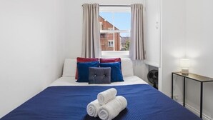 A neatly made bed with a blue bedspread, white pillows, and a grey accent pillow. There are two rolled towels on the bed. A small table with a lamp is next to the bed. - Well-equipped Stylish Apartment in Central Gloucester (Gloucestershire)