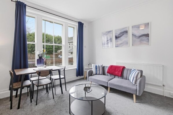 A modern living room with a gray sofa, a round glass coffee table, and a dining area with a wooden table and chairs.