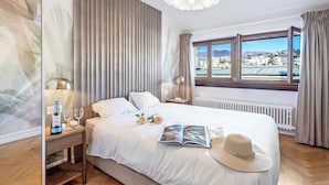 A hotel room with a large bed, a bedside table with a lamp, a window with a view of a cityscape, and a decorative hat on the bed. - Novos Arcadia 501 for 6 (Lugano)