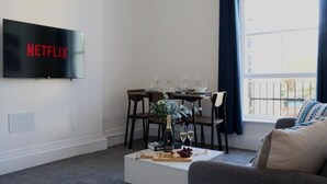 A living room with a TV displaying 'NETFLIX', a dining table with chairs, a coffee table with a bottle and glasses, and a sofa. - Well-appointed Stylish Apartment in Central Gloucester (Gloucestershire)