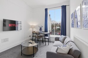 Un salon moderne doté d’un téléviseur à écran plat, d’une table basse ronde, d’un canapé, d’une table à manger avec des chaises et d’une grande fenêtre avec des rideaux.
