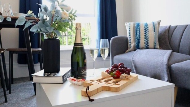 A living room with a white coffee table, a bottle of champagne, wine glasses, and a fruit platter. - Well-appointed Stylish Apartment in Central Gloucester (Gloucestershire)