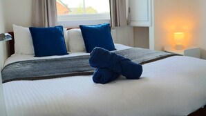 A neatly made bed with blue and white pillows, a folded towel, and a lamp on a bedside table. - Well-appointed Stylish Apartment in Central Gloucester (Gloucestershire)