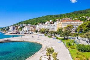 Paesaggio costiero con una passeggiata sul lungomare, edifici e una collina sullo sfondo.