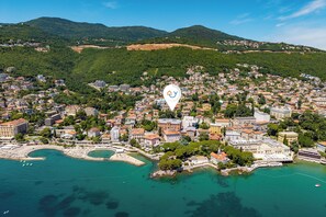 Una vista aerea di una cittadina costiera con un porto turistico, edifici residenziali fitti e un edificio bianco di rilievo con un logo blu.