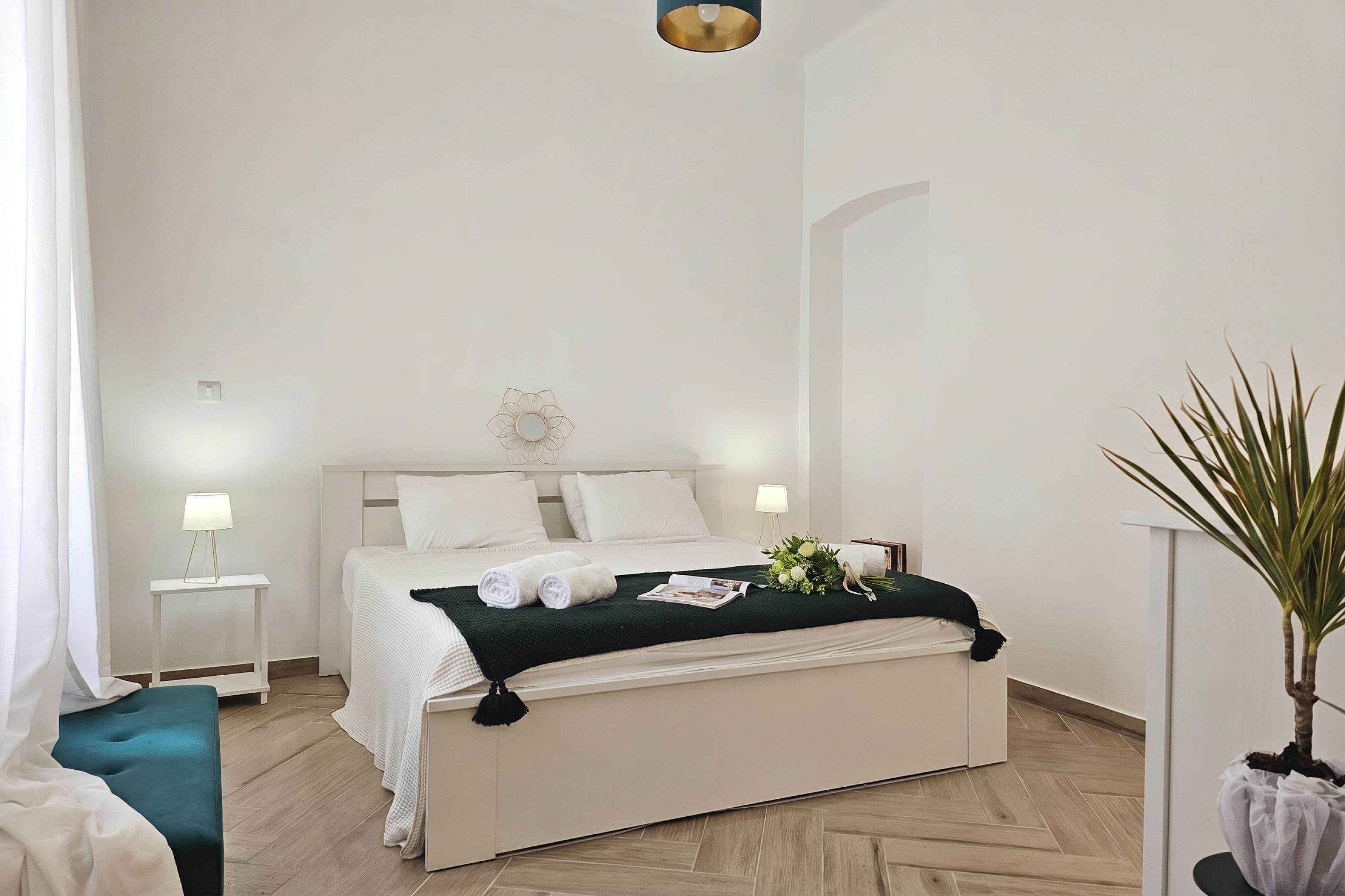 A hotel room with a bed, bedside tables, a bedside lamp, a green cushion, a plant, and a wall-mounted light fixture.