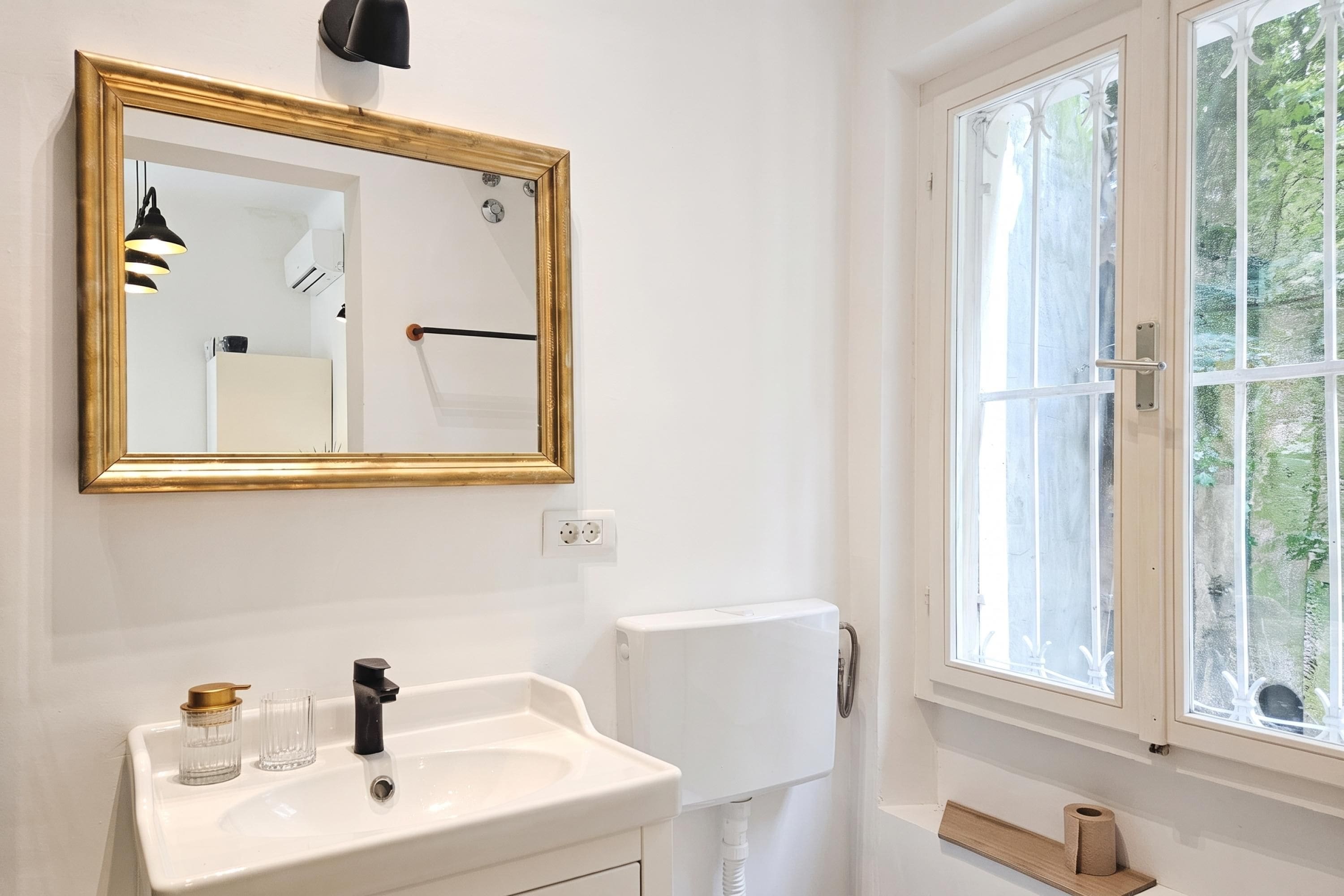 A bathroom with a white sink, a gold-framed mirror, a toilet, and a window with a view of greenery.