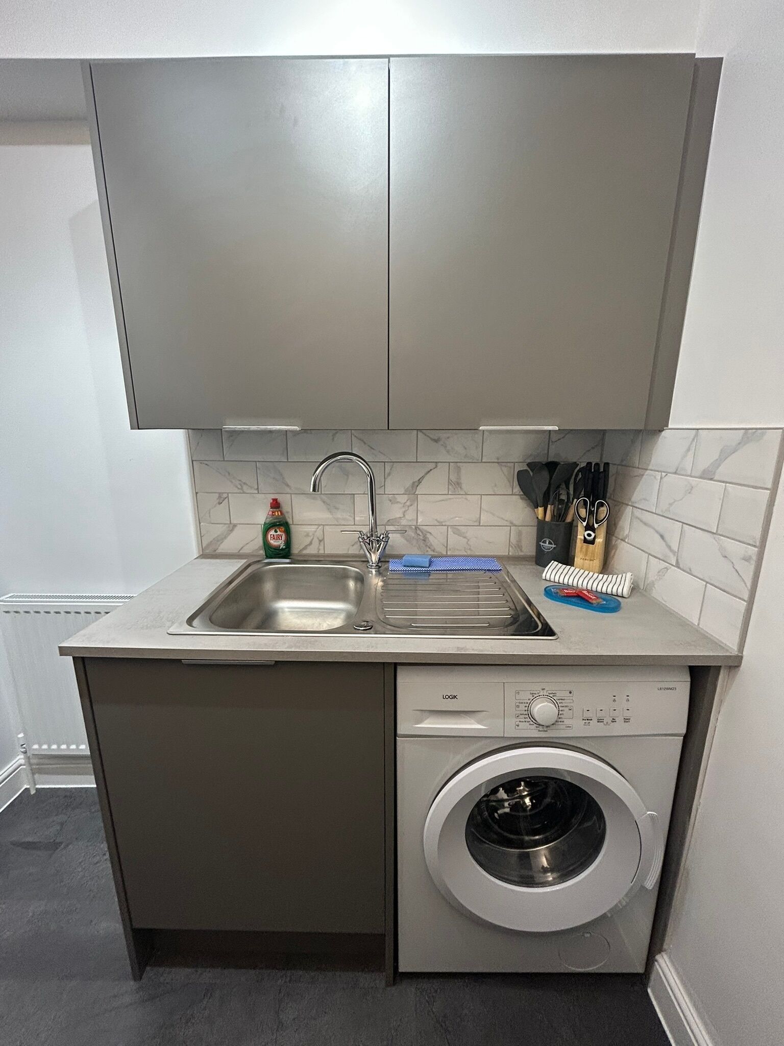 A compact kitchen with a built-in washing machine, a sink, and a countertop with a dish rack and utensils.