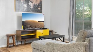 A modern living room with a large flat-screen TV mounted on a yellow and black cabinet, a wooden stool, a grey sofa, and a window with blinds. - Beautiful 1BD/1BA in University Hills jkxg (Austin)