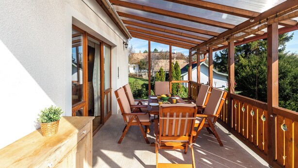 Eine überdachte Terrasse im Freien mit Holztisch und -stühlen, einem Korb mit Pflanzen und Blick auf einen Garten sowie Gebäude im Hintergrund.