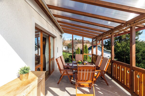 A covered outdoor patio with a wooden table and chairs, a basket with plants, and a view of a garden and buildings in the background.