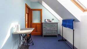 Ein kleiner Raum mit schrägem Dach, einer Holztür, einem Tisch mit Nähmaschine, einer Kommode und einem blauen Handtuch auf einem Ständer.
