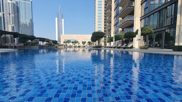 En swimmingpool med blå mosaikplattor, omgiven av moderna byggnader och en klar himmel.