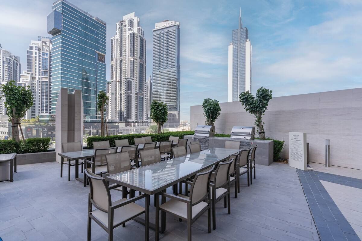 An outdoor dining area with a glass table and chairs, surrounded by a low wall and potted plants, with a city skyline in the background.