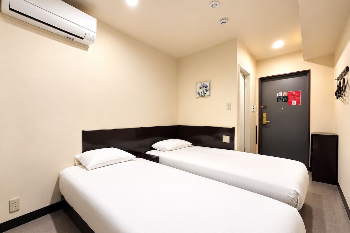 Ett hotellrum med två separata sängar, en mörk sänggavel, en luftkonditioneringsanläggning, en dörr med säkerhetslås och ett litet konstverk som hänger på väggen.