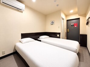Ett hotellrum med två separata sängar, en mörk sänggavel, en luftkonditioneringsanläggning, en dörr med säkerhetslås och ett litet konstverk som hänger på väggen.