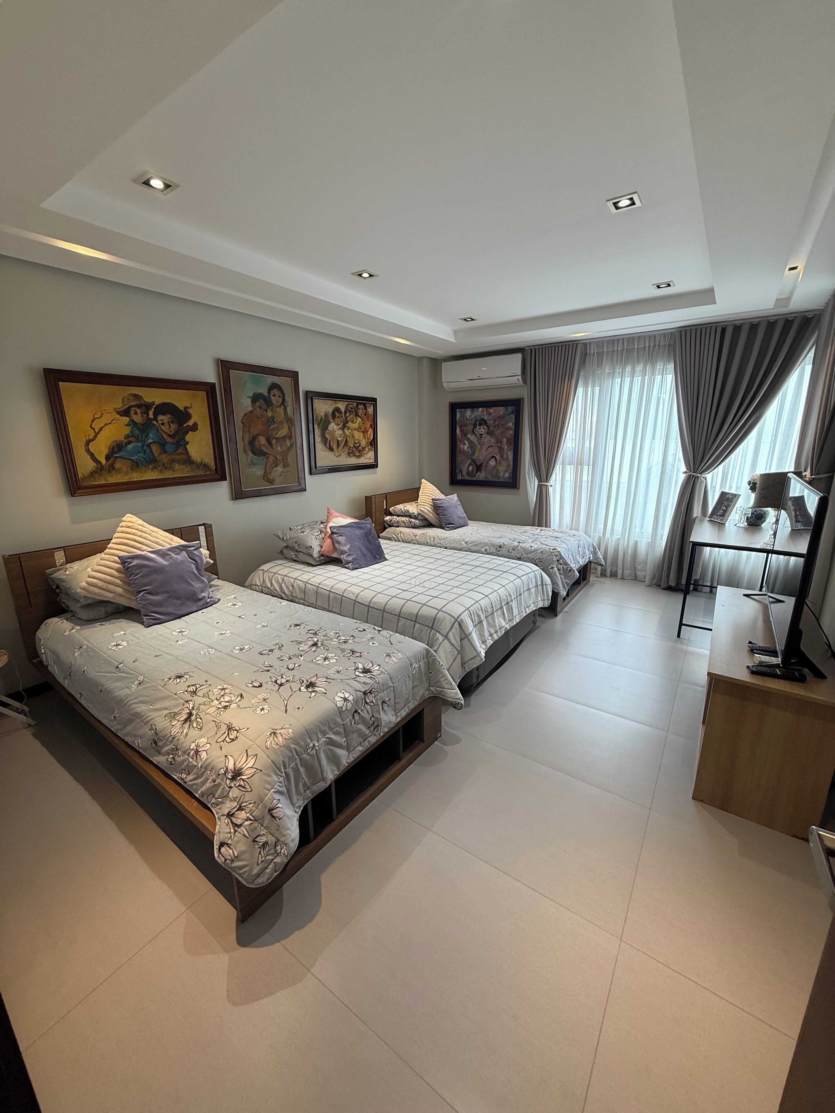 A bedroom with two beds, a desk, and a chair. There are paintings on the wall and an air conditioning unit.