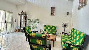 Un salón espacioso con techo alto, un candelabro, una mesa de centro de madera, dos sofás verdes con estampados amarillos, una planta en maceta y un ventilador.