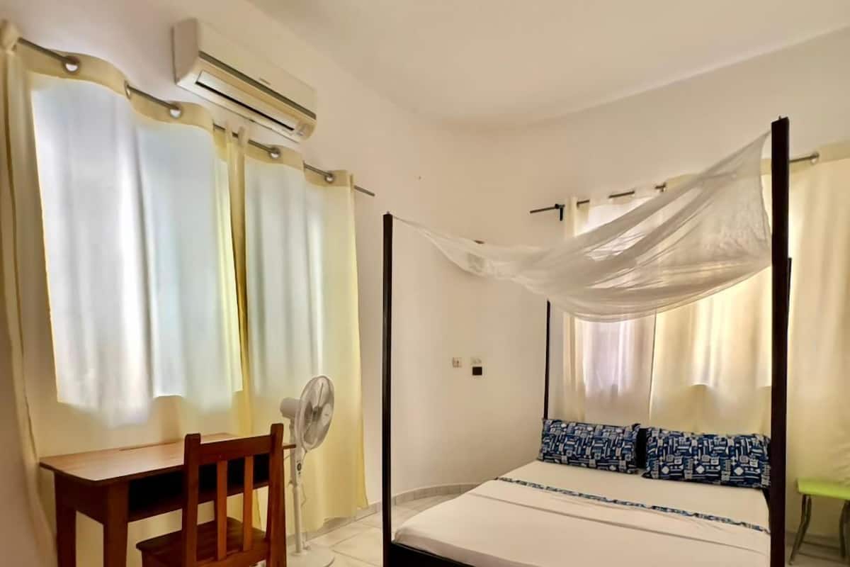 A bedroom with a four-poster bed, a desk with a chair, and a standing fan.