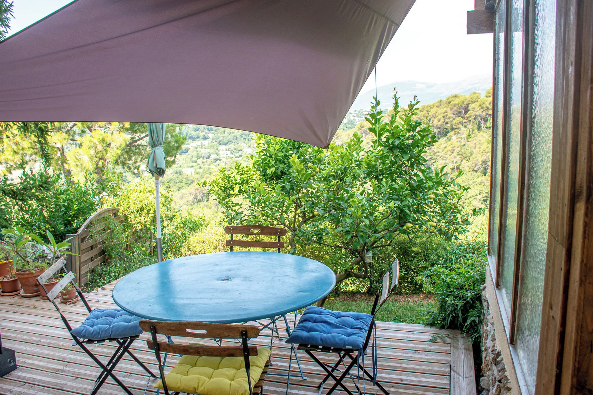 Un patio al aire libre con una mesa redonda y sillas, rodeado de plantas en macetas y vistas a un paisaje exuberante.