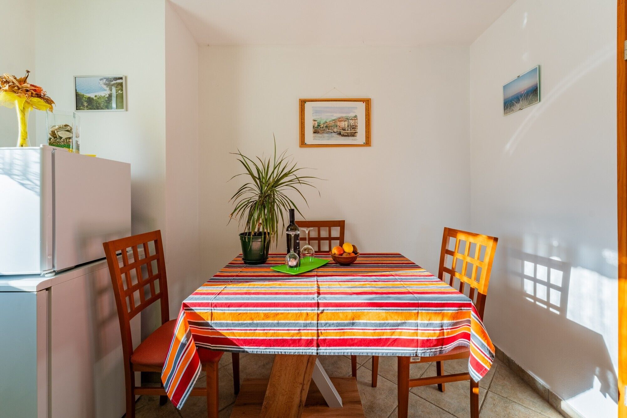 A dining area with a striped tablecloth, wooden chairs, a potted plant, and a small table with a bowl of fruit and a bottle.