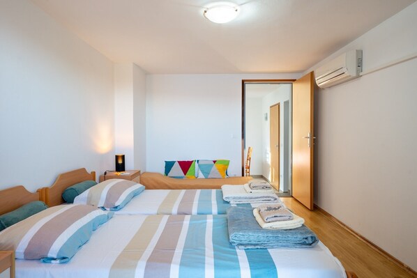 A hotel room with two beds, a bedside table with a lamp, a wall-mounted air conditioner, and a door leading to another room.