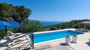 A rectangular swimming pool with a clear blue water, surrounded by lounge chairs and potted plants, overlooking a scenic view of the sea and hills.