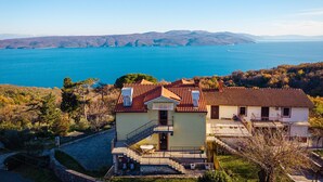 A coastal house with a balcony overlooking a bay, surrounded by trees and hills.