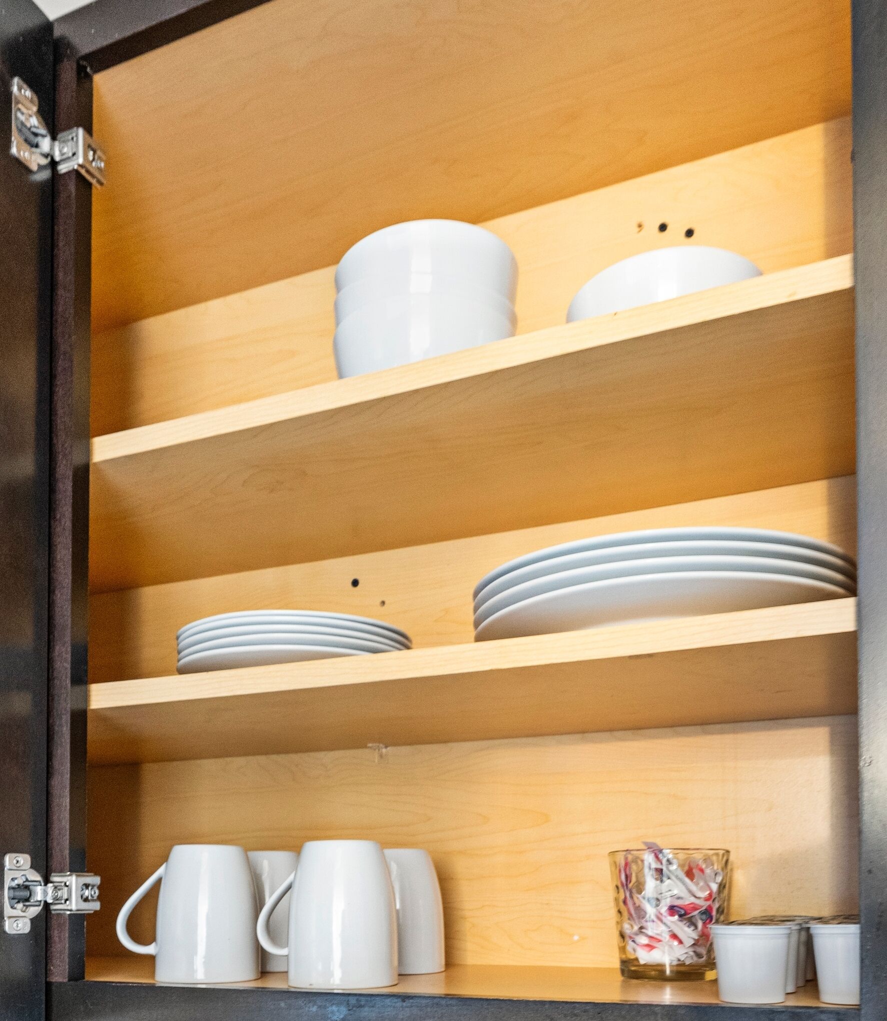 A wooden cabinet with shelves containing white mugs, plates, and a glass jar.