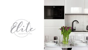 A modern kitchen with white cabinets, a black microwave, and a stainless steel sink. There is a vase with purple flowers on the counter. - Elite Apartments city center prestige (Gdansk)