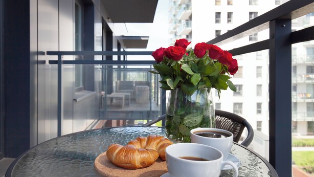 A balcony with a glass table, a vase of red roses, a croissant, and two cups of coffee. - Elite Apartments city center prestige (Gdansk)