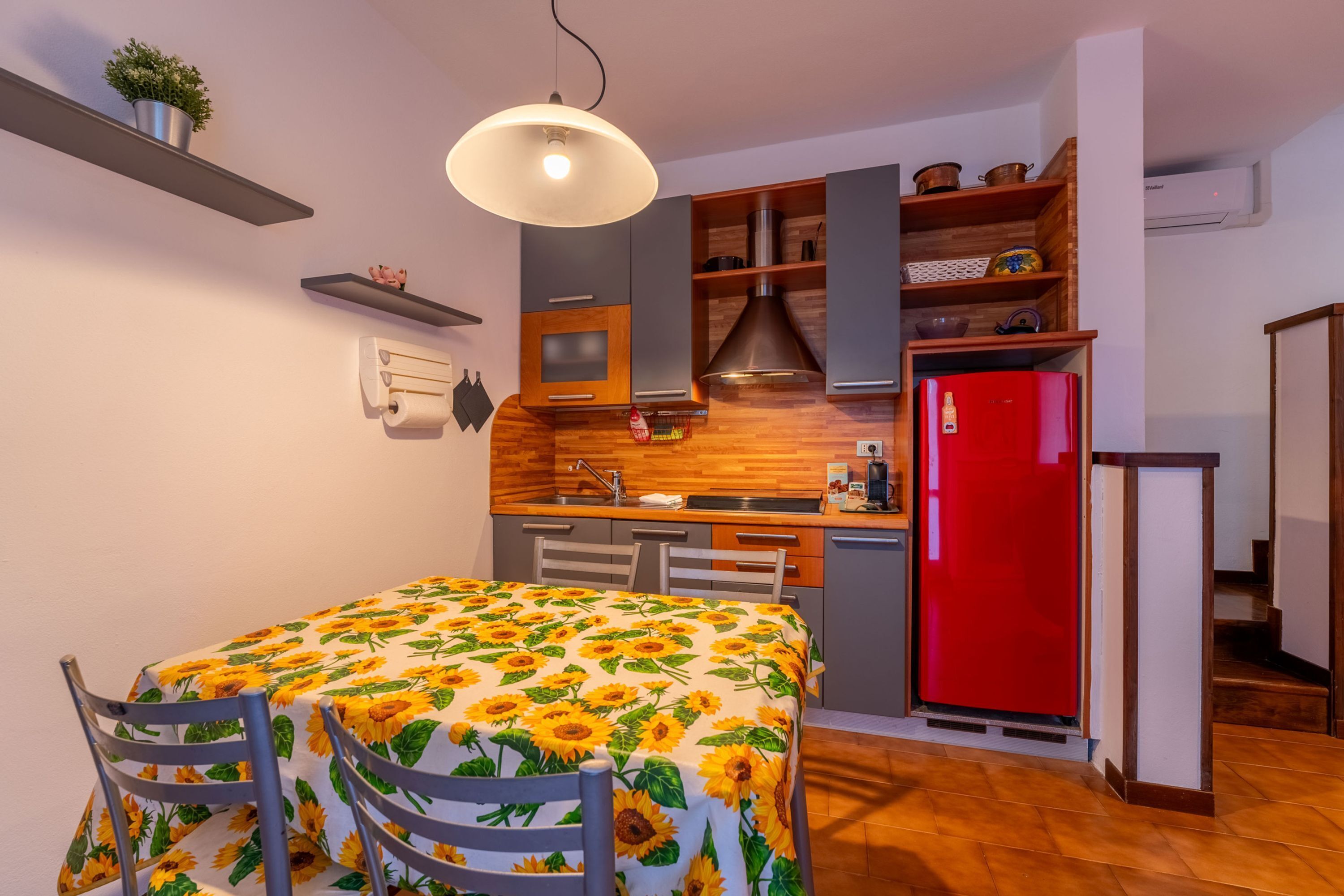 A compact kitchen with a wooden countertop, a red refrigerator, and a dining area with a table covered in a sunflower tablecloth.