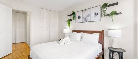 Quarto bem arrumado com uma cama grande, roupa de cama branca, cabeceira de madeira, mesa de cabeceira com abajur e prateleiras na parede com plantas.
