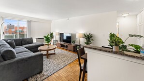 A modern living room with a sectional sofa, a round coffee table, a TV, and a dining area with a chair. - Gorgeous 2BR Next to BU y7k9 (Boston)