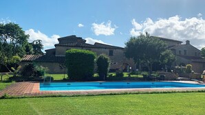 Una piscina con plataforma de ladrillo, rodeada de césped y árboles bien cuidados, y edificios de piedra al fondo.
