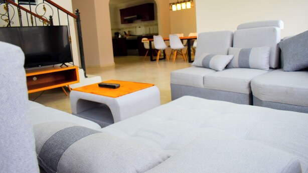 A modern living room with a grey sofa, a television, and a dining area in the background.