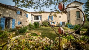 A rustic stone house with blue shutters, a garden with various plants, and a heart-shaped metal sculpture.