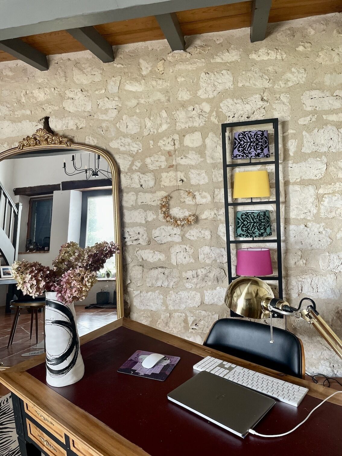 A room with a stone wall, a mirror, a desk with a laptop, a chair, and a lamp.