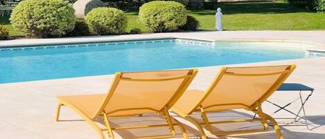 A poolside area with two yellow lounge chairs and a small table, surrounded by a well-maintained lawn and a building with blue shutters.