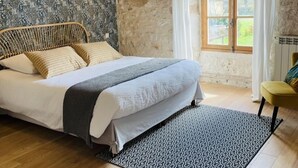 A bedroom with a bed, a patterned rug, wooden flooring, a stone wall, and a window with curtains.
